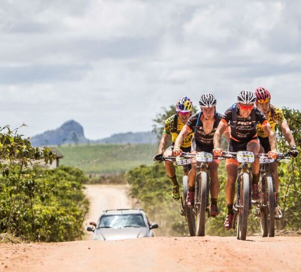 Ciclista de mountain bike em floresta brasileira, usando capacete e mochila, com vegetação densa e trilhas de terra ao redor, outro ciclista ao fundo.