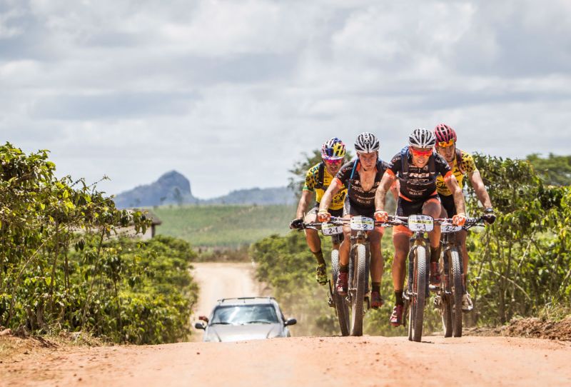 Ciclista de mountain bike em floresta brasileira, usando capacete e mochila, com vegetação densa e trilhas de terra ao redor, outro ciclista ao fundo.